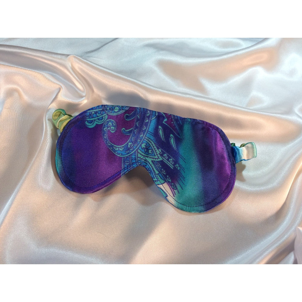 A beautiful turquoise blue and purple paisley print satin sleep mask laying on top of a white satin sheet.