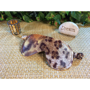 A brown leopard print sleep eye mask. Behind it is a lit candle and a stone with the word" dream" on it. Green plants are in the background.