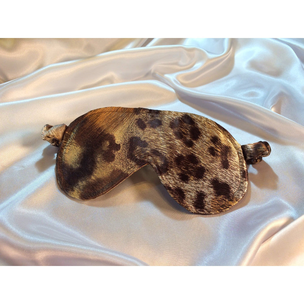 A brown leopard print satin eye mask. The sleep mask is lying on top of white satin sheets.