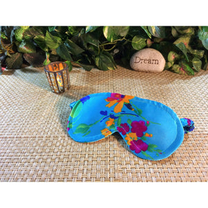 A  turquoise and floral print sleep mask . Behind it is a lit candle in a gold candle holder and a stone with the word "Dream". Various green plants are in the background.