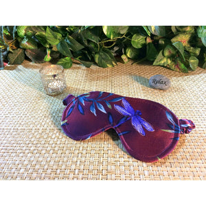 Maroon satin sleep eye mask with a blue dragonfly . There is a lit candle and a stone with the word "relax" behind it. In the background are various green plants.