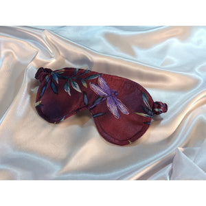 A maroon sleep eye mask with a blue dragonfly  on it. The mask is lying on top of a white satin sheet.