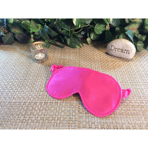 A bubblegum pink satin eye sleep mask with a candle burning behind it. A stone with the word "dream" is also in back of the sleep mask. Green plants are also behind.