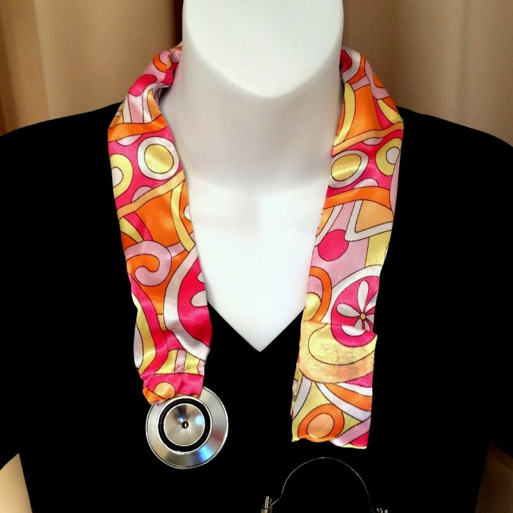 A stethoscope cover made from a yellow, pink and orange charmeuse satin print. The cover has a Velcro closing to keep it in place. The cover is hanging on a mannequin. 